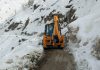 A JCB machine engaged in snow clearance operation on a road in Kargil district.